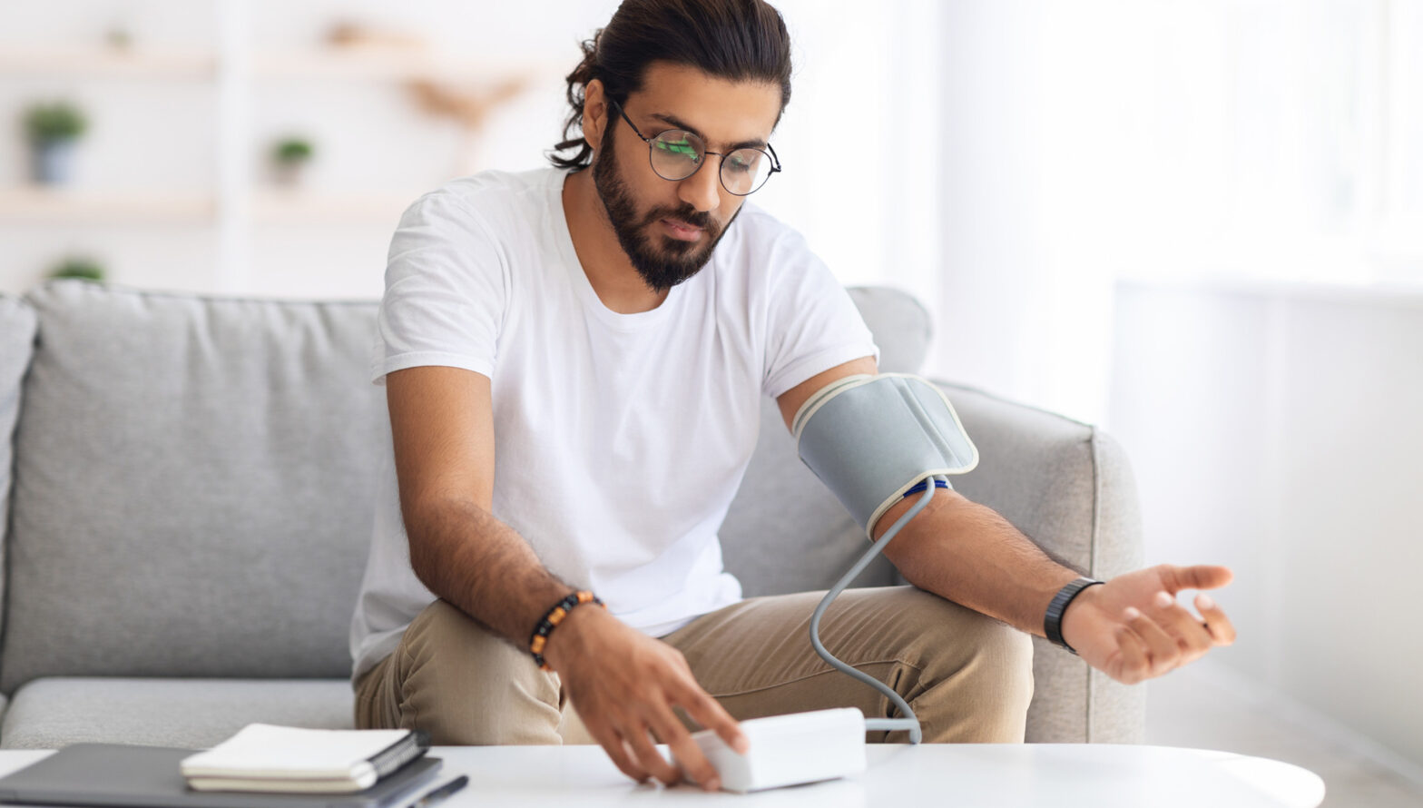 Young arab man sitting on couch, checking blood pressure and taking notes at home. Indian guy in white t-shirt using modern tonometer, experiencing hypertension caused by stress, copy space