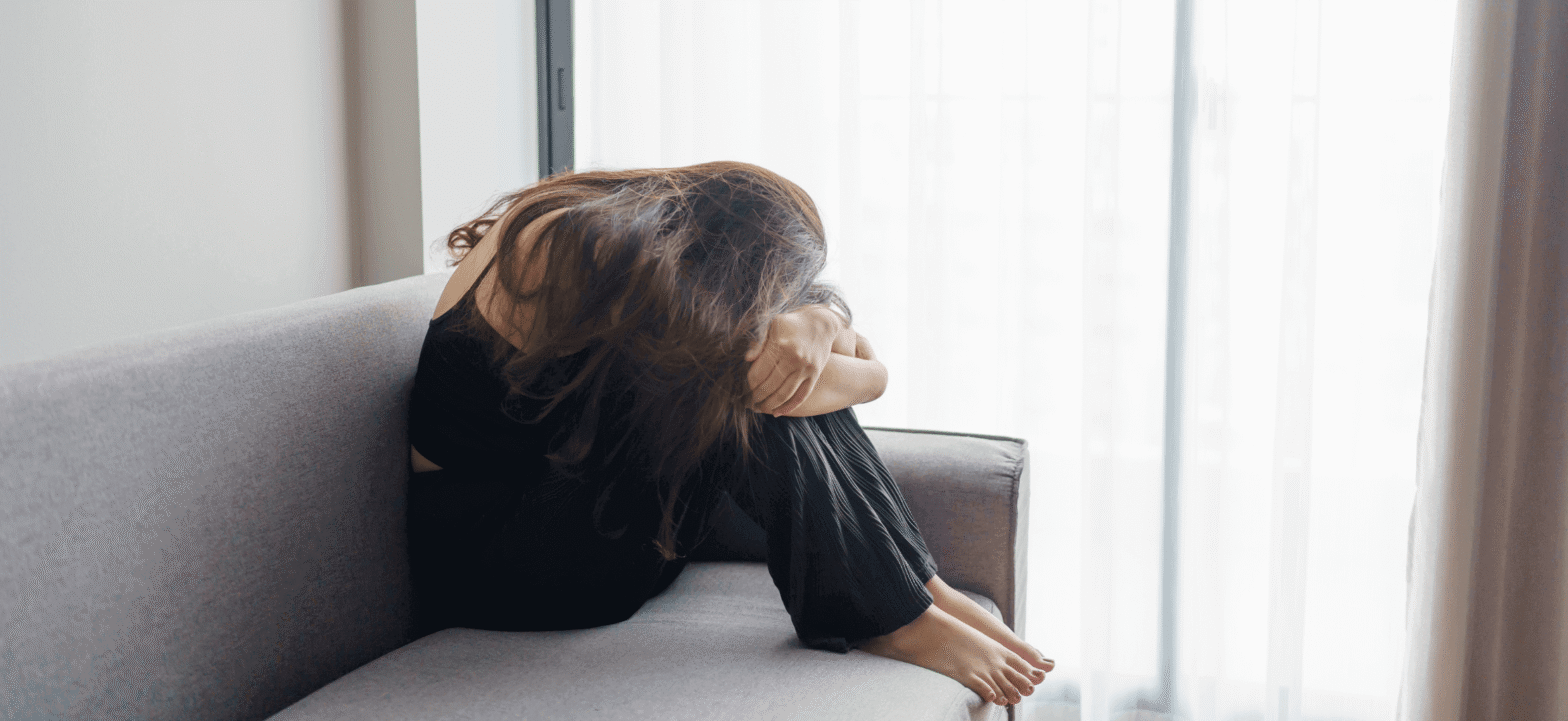 women wearing black with her knees pushed to her chest and her head down, siting on a couch in front of a sliding glass door with white curtains