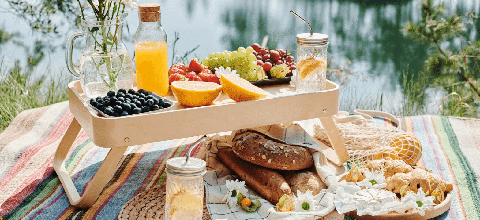 a luxurious picnic containing berries, bread, scones, juice, and a vase of flowers displayed on a woven blanket next to a body of water