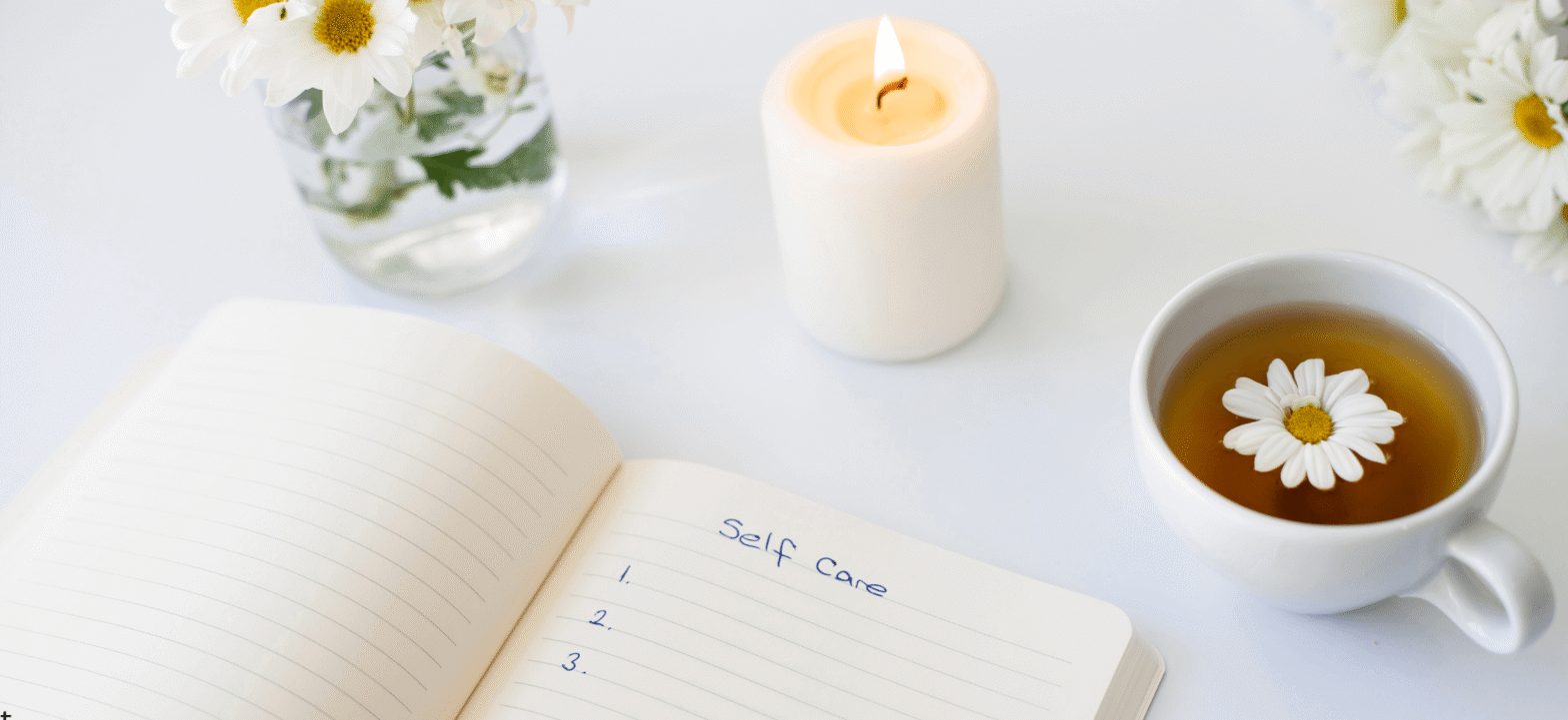 self care journal with relaxing setting created with a lit candle, vase of white flowers, and a cup of tea